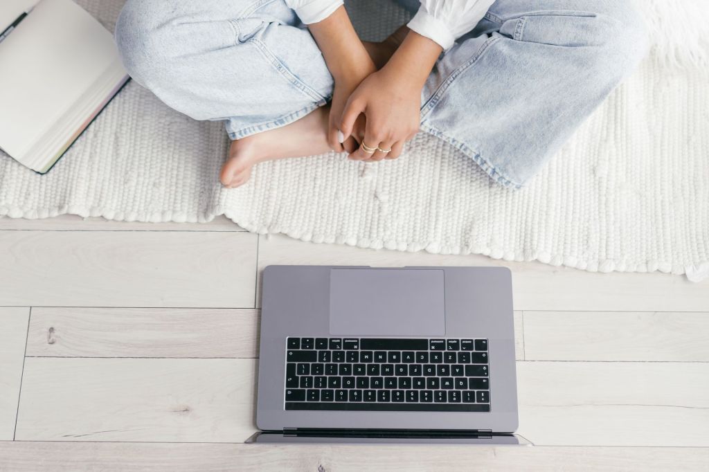 Top-down image of a barefoot person wearing pale denim and rings on their fingers sitting cross-legged on a white woven rug over a wood floor. In front of them sits an open laptop, and only the keyboard is visible. Image from Mikhail Nilov on Pexels.com.